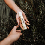 Hand holding Rosemary Leave-In Scalp Roller Serum among rosemary sprigs